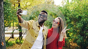 Beautiful couple African American man and Caucasian woman are taking selfie in park holding smartphone and posing