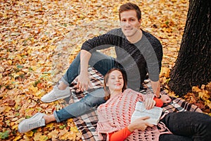 Beautiful couple is on blanket in autumn park. Young woman lying on guy`s legs. He sits on blanket. They look on caemera