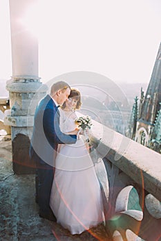Beautiful couple, bride and groom posing on old balcony with column, cityscape background