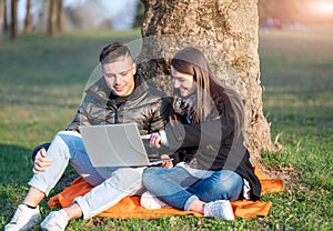 A beautiful couple in casual wear is having fun with the laptop outdoors. Sitting at the foot of a tree, you enjoy nature on a