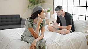 Beautiful couple disagreeing in their bedroom, sitting on the bed, expressing conflict while using a smartphone, a problem causing