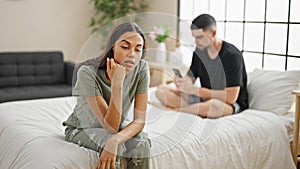 Beautiful couple disagreeing in their bedroom, sitting on the bed, expressing conflict while using a smartphone, a problem causing