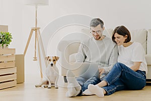 Beautiful couple with dog sit on floor in spacious living room, make purchasing online, buy furniture for new home, have to unpack