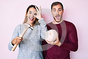 Beautiful couple holding piggy bank and hammer smiling happy doing ok sign with hand on eye looking through fingers