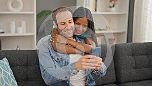 Beautiful couple hugging each other sitting on sofa using smartphone at home