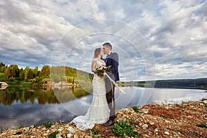 Beautiful couple in love kissing while standing on ground by lake. Wedding couple at sunset, blue cloud sky, love and tender feel