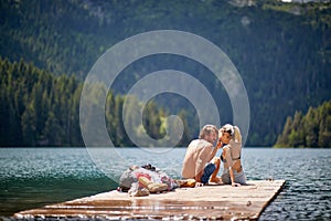 Beautiful couple in love sitting on wooden jetty by lake. Backpackers enjoying summer day in nature. Lifestyle, love, adventure,