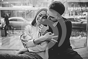 Beautiful couple. Loving guy hugs his charming girlfriend sitting on the windowsill in a cozy cafe. Black and white