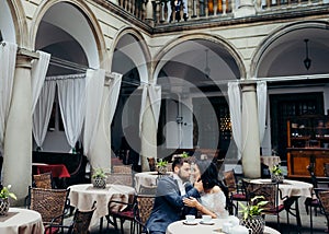 Beautiful couple of newlyweds is sitting at the table in the outdoor restaursnt. The handsome groom is softly stroking