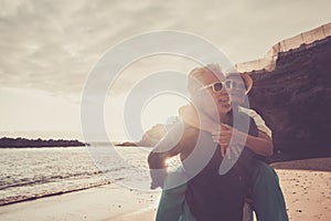 Beautiful couple of seniors at the beach having fun together and playing - mature man carry his wife woman with love and forever