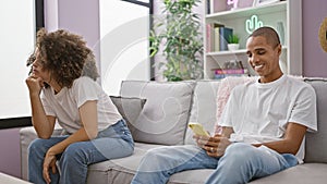 Beautiful couple sitting at home, unhappy on a roomy sofa while confident boyfriend happily engages with smartphone, problem