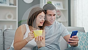 Beautiful couple sitting on sofa together using smartphone drinking coffee at home