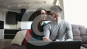 Beautiful couple sitting on a sofa, young man checking his emails in laptop and talking to girlfriend , kissing her on