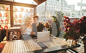 Beautiful couple sitting at sunset on terrace in city cafe with laptop on table, hugging and talking. Love story portrait of a