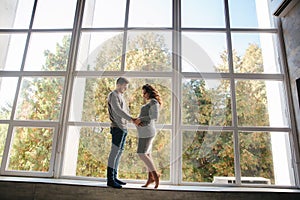 Beautiful couple stand in front of big window in studio. Pregnant woman with her husband