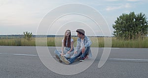 Beautiful couple of teens sitting on skateboard on road and having romantic talk