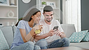 Beautiful couple together in love, confidently smiling, texting on smartphone over coffee on living room sofa at home