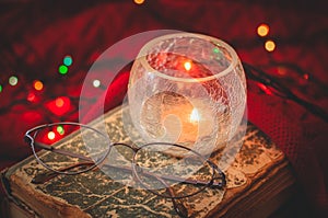 Beautiful cozy christmas winter scene with a book, eyeglasses, a burning candle and glowing xmas garland close-up