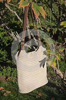 Beautiful cream crocheted handbag with brown handle and nature leaves behind the bush in the garden in summer