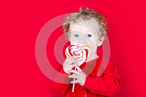 Beautiful curly baby girl eating a heart shaped candy on red background