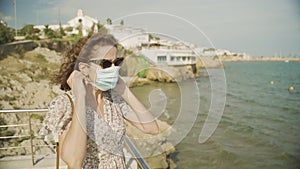 Beautiful curly brunette tourist puts on a surgical mask during Covid-19 with the Mediterranean village of Sitges on the