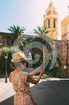 Beautiful curly brunette tourist with a surgical mask taking a selfie in the Mediterranean village of Sitges, spain.