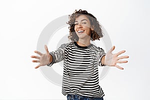 Beautiful curly girl reaching hands, stretching arms to hug, receiving smth, standing in blouse against white background
