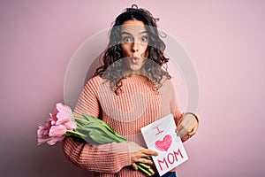 Beautiful curly hair woman holding love mom message and tulips celebrating mothers day scared in shock with a surprise face,
