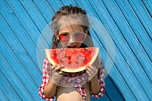 A beautiful curly-haired girl in a plaid shirt bites a large piece of watermelon