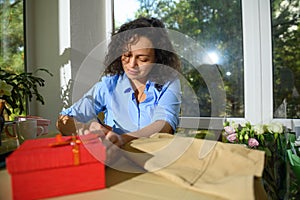 Beautiful curly haired Latin American woman taking notes in a floral workshop. Beige apron and red gift box on the table