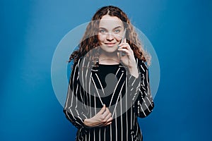 Beautiful curly young girl in casual wear looking at the camera speaks by phone while standing isolated over blue background