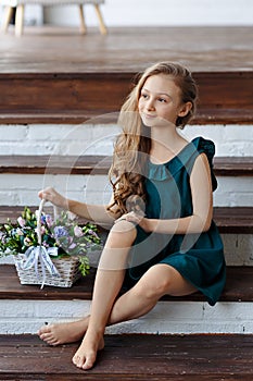 Beautiful cute baby girl with long hair in a green dress sits in a bright studio and holds a basket with natural flowers in her