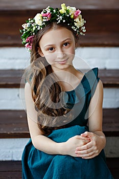 Beautiful cute baby girl with long hair in a green dress sits in a wreath of fresh flowers on her head and looks at the camera