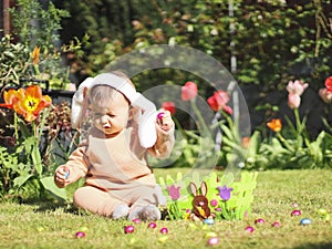 Beautiful and cute caucasian baby girl with headband bunny ears sits on the lawn with flowers.
