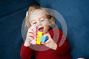 Beautiful cute caucasian baby girl, toddler with curly blond hair playing with erector set, lying on sofa,smiling,kid