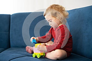 Beautiful cute caucasian baby girl, toddler with curly blond hair playing with erector set, sitting on sofa, serious