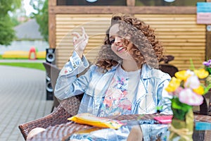 Beautiful cute cheerful girl in a bright dress with curly hair and bright make-up sitting at an outdoor cafe and waiting for