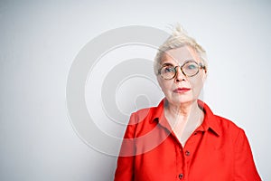 Beautiful cute elderly woman in glasses and in a red shirt looking at the camera while standing on a gray background