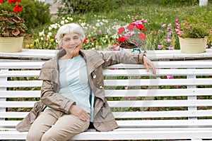 Beautiful cute elderly woman sitting on a park bench with flowers.