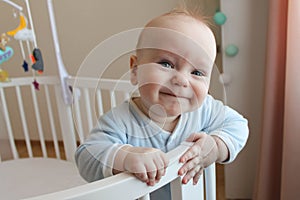 A beautiful cute European baby is standing in a white oval four-poster bed and smiling.