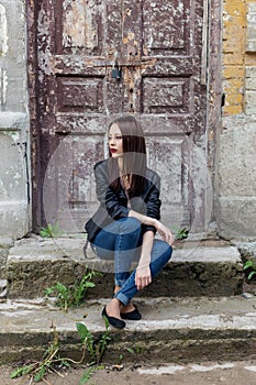 Beautiful cute fashion girl with dark hair with sunglasses in a leather black jacket sitting on the stairs the porch of old