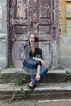 Beautiful cute fashion girl with dark hair with sunglasses in a leather black jacket sitting on the stairs the porch of old