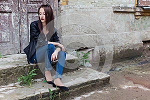 Beautiful cute fashion girl with dark hair with sunglasses in a leather black jacket sitting on the stairs the porch of old