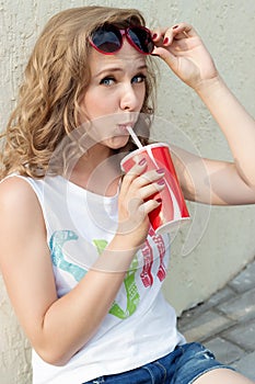 Beautiful cute girl with curly hair in sunglasses with a paper glass and a straw drinking a drink in the city on a sunny summer ev