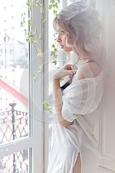 Beautiful cute girl near the window in the morning in a white shirt with a cup of coffee