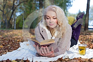 Beautiful cute happy smiling girl lying on the ground and reads a book in autumn Park the Park with a mug of hot tea