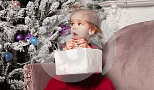 A beautiful cute little girl dressed in an elegant evening red dress sits on the couch and opens a New Year`s gift.