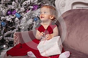 A beautiful cute little girl dressed in an elegant evening red dress sits on the couch and opens a New Year`s gift.