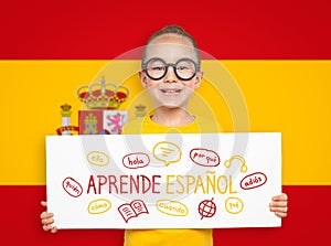 Beautiful cute little girl holding a white board with Learn Spanish text in Spanish and illustrations in front of blurry flag of S