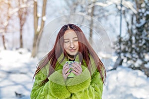 Beautiful cute pretty redhead female teenager in a green fur coat drinking coffee in the sunny winter city park.